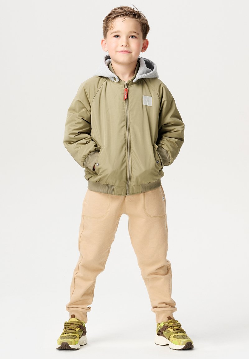 Olivgrüne Bomberjacke mit grauem Kapuzenumschlag, kombiniert mit hellbeigen Jogginghosen und gemusterten Sneakers in Gelb, Braun und Weiß.