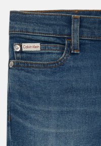 Jeans en denim Calvin Klein de couleur bleu foncé. Présentent un design classique, des coutures orange et une petite étiquette placée près de la poche avant.