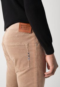 Pantaloni din corduroy bej cu dungi orizontale vizibile, o etichetă din piele pe spate și trei accente cusute albastre pe buzunar.
