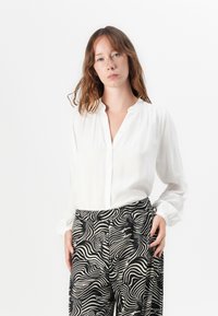 White button-up blouse with long sleeves, paired with high-waisted black and cream patterned trousers featuring a wavy design.