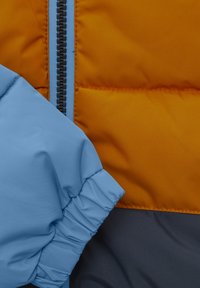 Isolerad jacka med en orange kropp och en blå ärm, grå detaljer och en mittdragkedja. Materialet verkar slätt och glänsande.