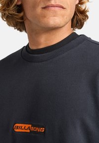 Sudadera de cuello redondo en color navy, fabricada en un material suave, con un parche texturizado en negro que presenta un logo naranja de "Billabong" en el lado izquierdo del pecho.