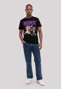 Black cotton t-shirt featuring bold purple "BIEBER" text and photographic prints of a male figure. Paired with blue denim jeans and white sneakers.