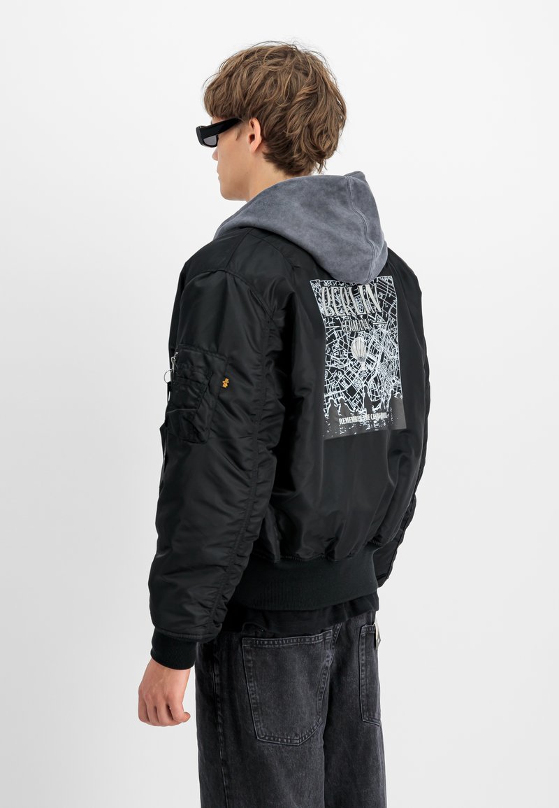 Chaqueta bomber negra con capucha gris, puños elásticos y diseño gráfico en la parte posterior. Incluye un bolsillo con cremallera en la manga y una textura suave y brillante.