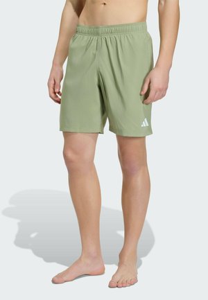 Mężczyzna w jasnozielonych sportowych krótkich spodenkach z logo Adidas, stojący boso na jasnym tle.