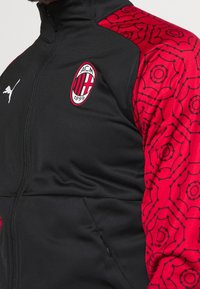 Black athletic jacket with red patterned sleeves. Features a Puma logo on the chest and an AC Milan emblem. Smooth texture, zippered front.