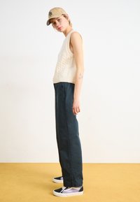 Un top fără mâneci tricotat cu cablu de crem, pantaloni inchisi la culoare și adidași de tip low-top negri și mov, purtând o șapcă bej cu un logo. Fundal galben.