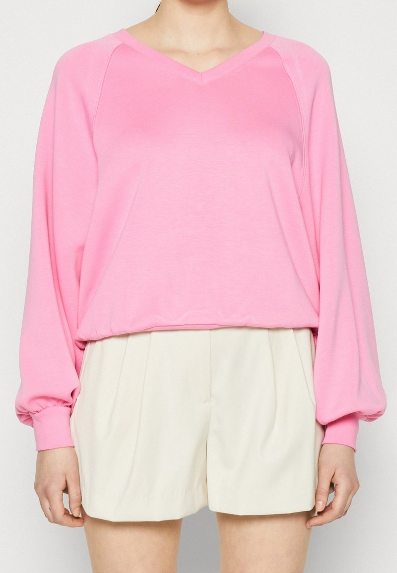 Personne portant un haut rose ample à manches longues avec des manches retroussées et un short beige plissé taille haute, debout devant un fond uni.