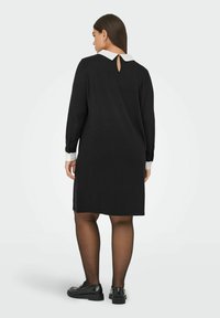 �Žena u crnoj haljini do koljena s bijelom kragnom i manžetama, crnim prozirnim čarapama i crnim loafericama, stoji okrenuta leđima.