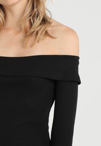 Černý top s odkrytými rameny, s žebrovanou texturou a dlouhými rukávy, se širokým přehnutým výstřihem a hladkým materiálem.