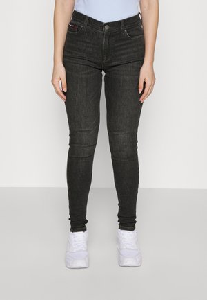 Person iført sorte skinny jeans og hvide sneakers stående mod en ensfarvet lys baggrund.