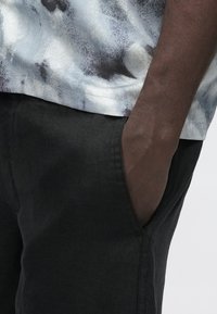 Camisa de manga curta com padrão abstrato em azul claro e cinza, combinada com calças pretas; bolso visível com detalhe em tecido texturizado.