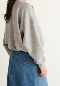 Šedá oversized mikina s kulatým výstřihem, v kombinaci s modrou džínovou sukní. Materiál vypadá měkce a má hladký povrch.