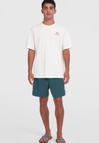 Light beige cotton t-shirt with a green logo detail, paired with teal swim shorts and blue patterned sandals, on a neutral background.