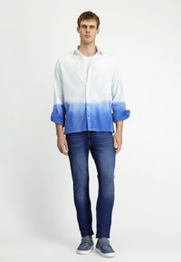 Knopjesoverhemd met een wit-blauwe gradient, gestructureerde stof, opgerolde mouwen; gecombineerd met donkere jeans en blauwe instappers.