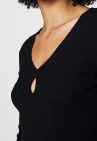 Schwarzes geripptes Oberteil mit V-Ausschnitt und Schlüssellochdetail. Der Stoff ist glatt und enganliegend, wodurch die Körperform betont wird.