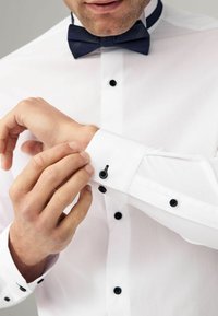 Camisa de vestir blanca con una textura suave, que cuenta con botones negros y un puño abotonado. Una pajarita azul marino añade contraste al cuello.