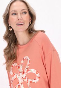 Rosa sweatshirt med en blommig applikation i vitt och rött. Modellen bär cirkulära örhängen med färgglada accenter. Mjuk tygkvalitet och avslappnad passform.