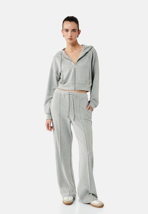 Frau trägt hellgrauen Kapuzenpullover mit Reißverschluss und passende weite Jogginghosen mit weißen Sneakern, steht vor schlichtem Hintergrund.