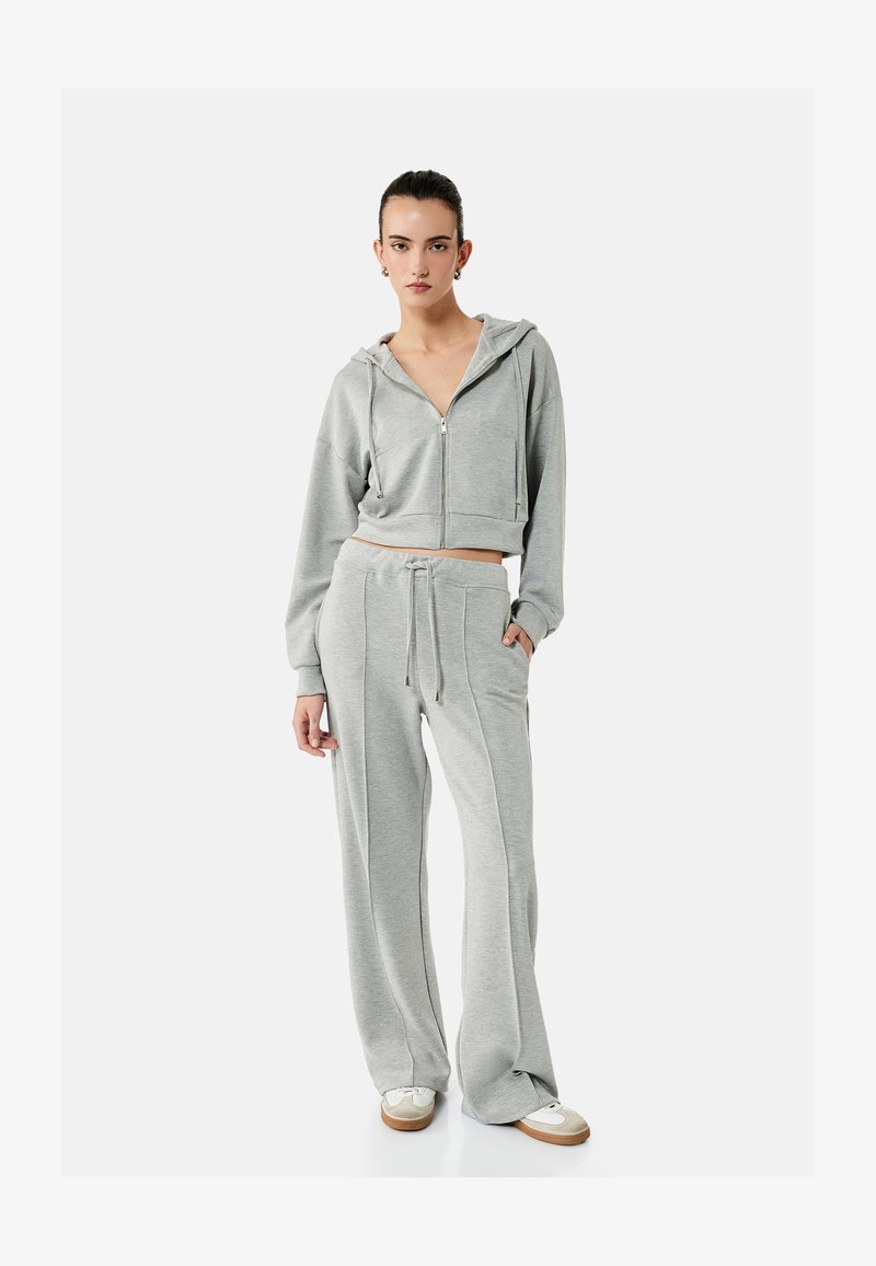 Kobieta w jasnoszarej bluzie z kapturem na zamek i dopasowanych szerokich spodniach dresowych, białe trampki, stojąca na jednolitym tle.