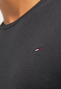 Närbild av en person som bär en svart t-shirt med en liten broderad röd, vit och blå rektangulär logotyp på bröstet.