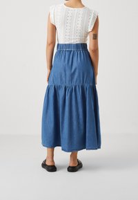 Jeansmaxikjol med insamlad midja, lagerdesign och subtil textur i en medelblå nyans. Matchas med en vit broderad topp.