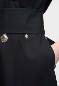 Vêtement noir avec une large ceinture et des charms dorés en forme de coquillages suspendus le long de la taille, main visible dans la poche latérale droite.