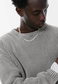 Grå, ribbad tröja med vid halsringning och långa ärmar. Har ett silverkedjearmband som framhäver textur och lagerdesign.