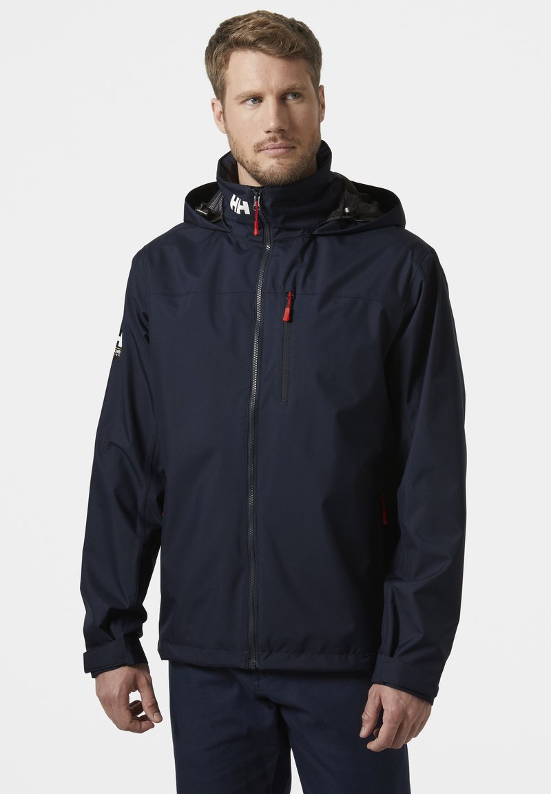 Navyblauwe waterdichte jas met een capuchon, ritssluiting aan de voorkant, rode ritsaccenten, verstelbare manchetten en logo op de schouder.