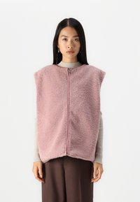 Vesta pufo roz, cu guler înalt și fermoar în față, având o textură moale și un design supradimensionat, purtată peste un pulover cu model nervurat.