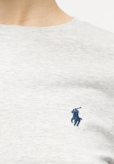 Κοντινό πλάνο ενός ανοιχτό γκρι t-shirt με στρογγυλή λαιμόκοψη και ένα μικρό σκούρο μπλε κεντητό λογότυπο παίκτη πόλο στο στήθος.