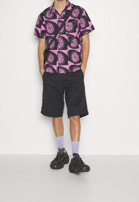 Lyhythihainen nappikauluspaita vaaleanpunaisena ja mustana, jossa on spiraalimalli, yhdistetty mustiin shortsihousuihin ja mustiin urheilukenkiin, jotka on korostettu laventelinvärisillä sukilla.