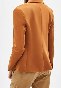 Blazer orange sur mesure avec un design structuré, featuring un col à revers cranté, une texture lisse et une silhouette cintrée.