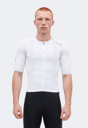 Ung mand med kort rødt hår iført en tætsiddende hvid cykeltrøje med korte ærmer og sorte cykelshorts, stående mod en ensfarvet baggrund.