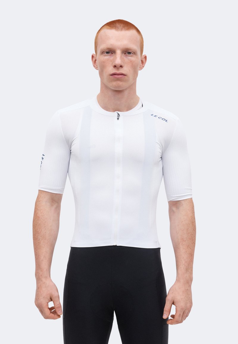 Junger Mann mit kurzen roten Haaren, der ein enganliegendes weißes Radtrikot mit kurzen Ärmeln und schwarze Radhosen trägt, steht vor einem einfarbigen Hintergrund.