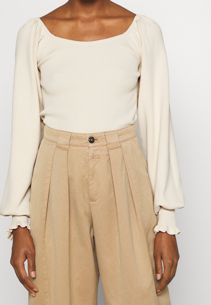 Femme portant un haut côtelé crème à manches longues bouffantes et un pantalon plissé beige taille haute avec une fermeture à boutons.