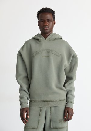 Giovane uomo che indossa una felpa oversize verde tenue e pantaloni coordinati con cuciture visibili e logo Alpha Industries sul davanti della felpa.