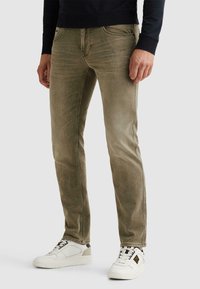 Hombre con vaqueros ajustados de color verde oliva deslavado y zapatillas blancas, de pie frente a un fondo claro y liso.