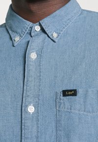 Camicia in denim blu chiaro con bottoni, collo a camicia, bottoni bianchi e una piccola etichetta nera con la scritta "Lee" in oro sulla tasca.