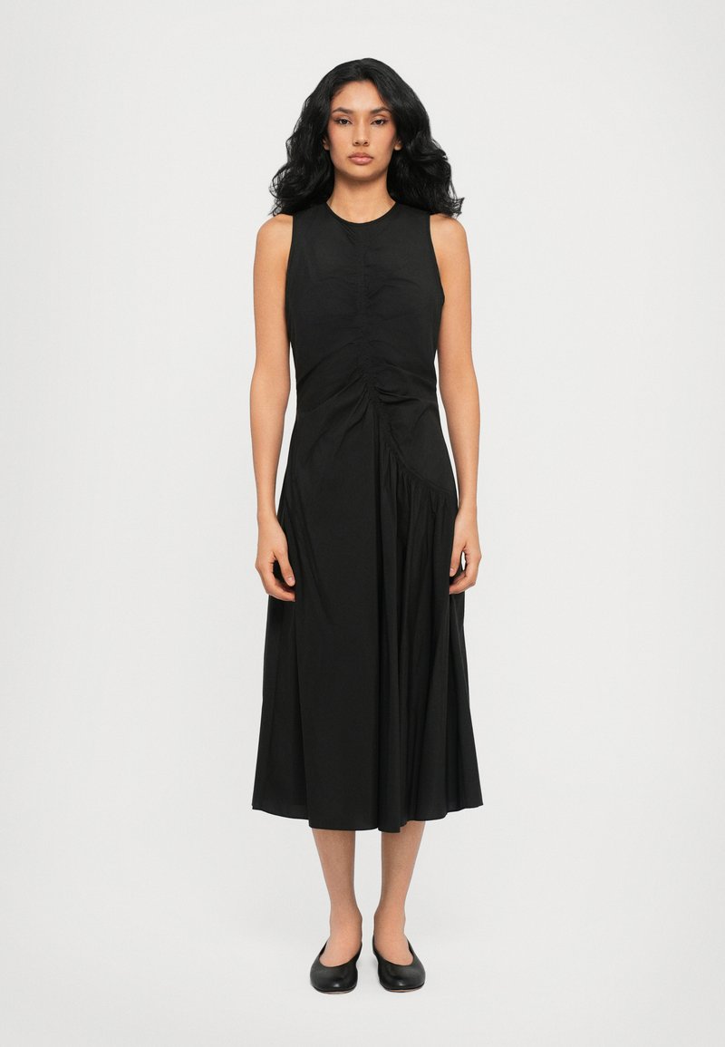 Black sleeveless dress with gathered front detail, flared skirt, and smooth texture. Paired with simple black flats.