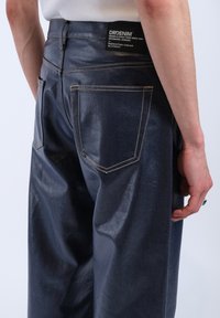 Dark blue denim jeans featuring back pockets with contrasting orange stitching and a black label near the waistband. Smooth texture.