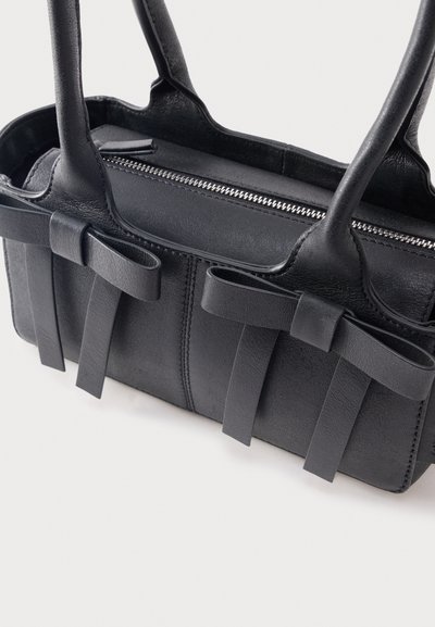 Μαύρη δερμάτινη τσάντα χειρός με δύο λαβές, φερμουάρ στην κορυφή και διακοσμητικούς δερμάτινους φιόγκους στις μπροστινές πλευρές.