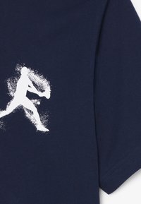 Silueta de una persona balanceando una raqueta de tenis con una pelota cerca, en blanco sobre un fondo de tela azul marino, probablemente una camiseta.