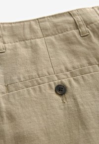 Pantalons beiges en tissu de lin texturé, avec une poche arrière et un bouton noir de fermeture. Design simple sans motifs supplémentaires.