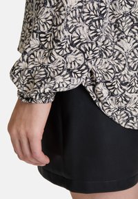 Schwarz-weiße Langarmbluse mit floralem Muster, leichtem Stoff und gerafften Ärmeln. Kombiniert mit einem schwarzen Rock.