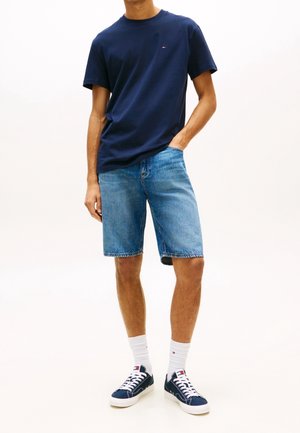 Homme portant un t-shirt bleu marine, un short en jean bleu clair, des chaussettes blanches et des baskets bleu marine, debout avec une main dans la poche.