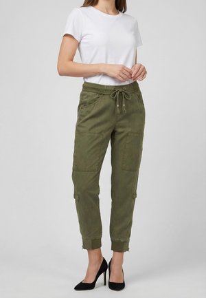 Mujer con pantalones cargo verde oliva con cintura con cordón, camiseta blanca de manga corta y zapatos de tacón negros, de pie frente a un fondo gris.