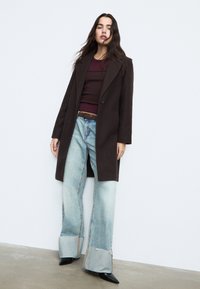 Hnědý kabát z vlněné směsi přes vínově červený top, doplněný širokými světle modrými džíny s roztřepenými lemy a černými koženými špičatými botami.