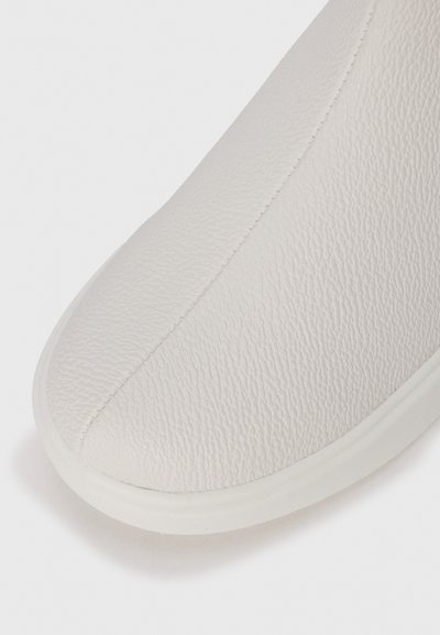 Vita lädersko med en texturerad yta, rund tå och kontrasterande vit gummisula. Sömsömmar synliga längs sidan.