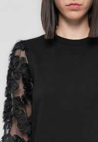 Black sweatshirt with a rounded neckline, featuring sheer black mesh sleeves adorned with layered floral appliqués. Smooth fabric texture.